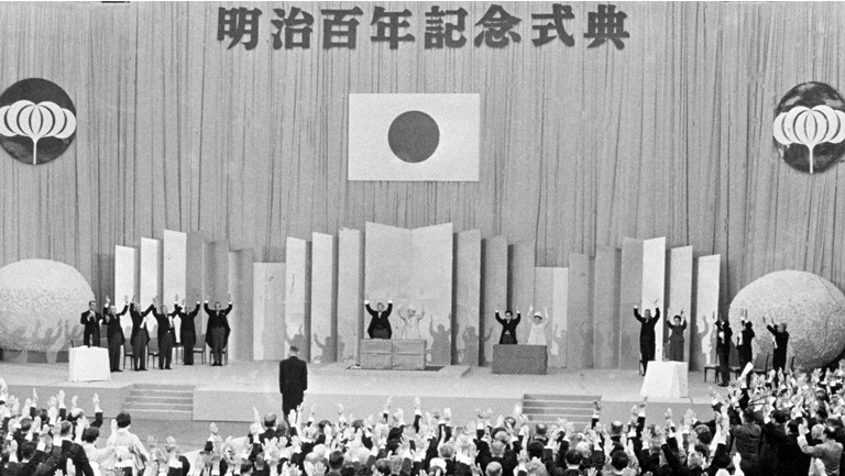 Prime Minister Eisaku Sato leads the hall in shouts of banzai at a grand ceremony commemorating the Meiji centennial, held on October 23, 1968, at the Budokan in Tokyo. At center stage are the former emperor and empress. ©Kyodo News